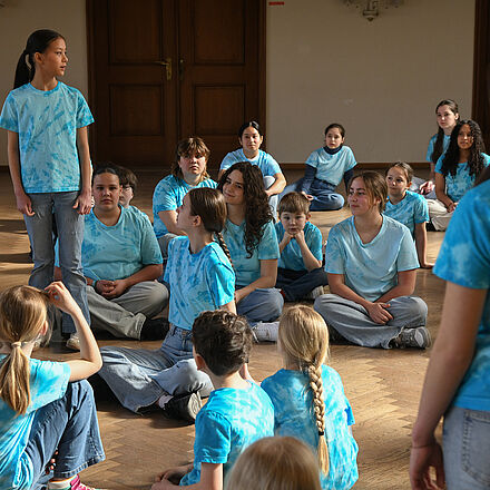 Der Kinderchor sitzt auf dem Boden. Zwei Mädchen stehen dazwischen. Alle tragen ein blaues Oberteil.