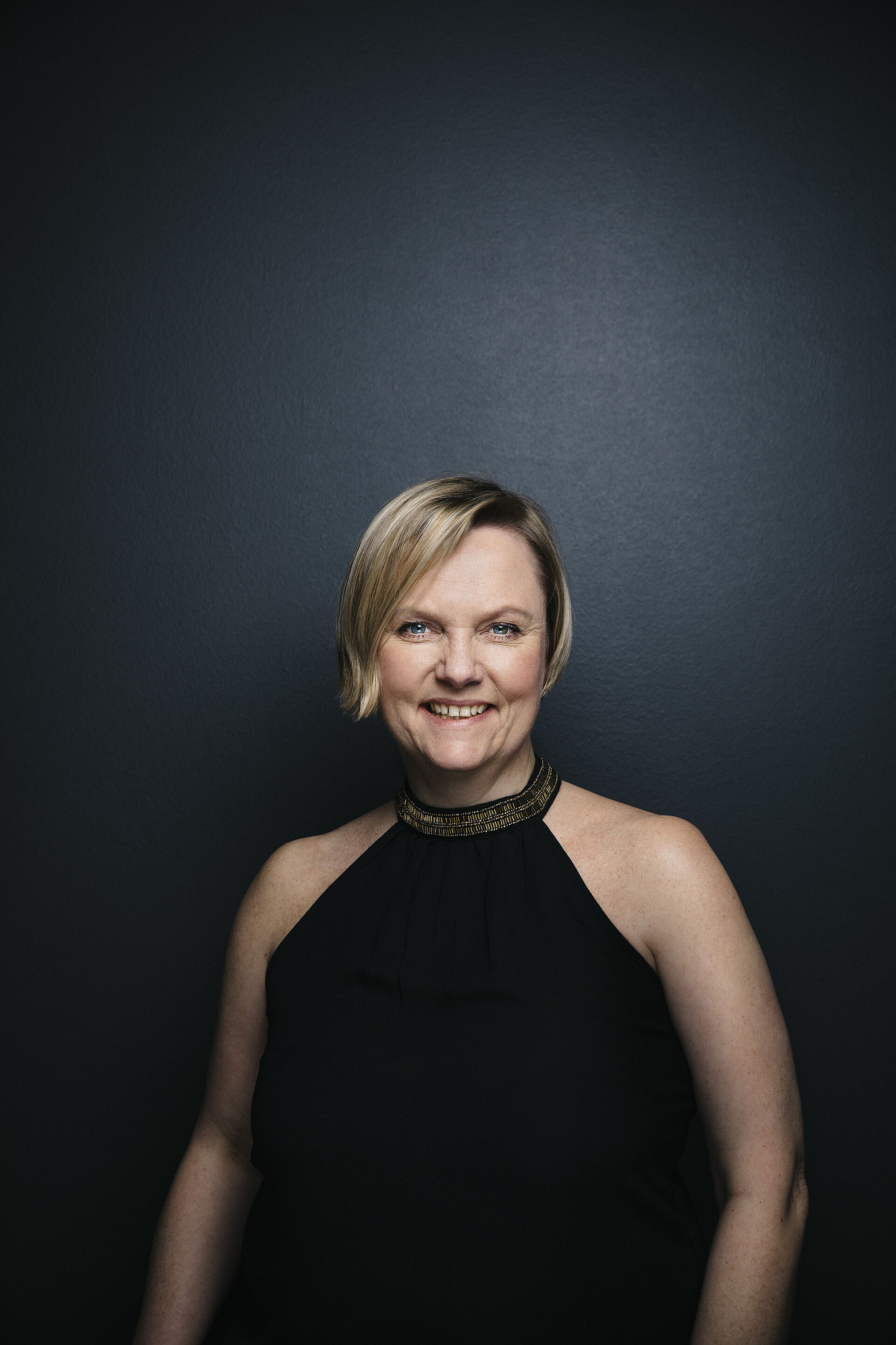 Portraitfoto von Sabine Arthold. Sie steht grinsend vor einer grauen angeleuchteten Wand. Sie trägt ein schwarzes schulterfreies Oberteil. Ihr blondes kurzes Haar umspielt ihr Gesicht.