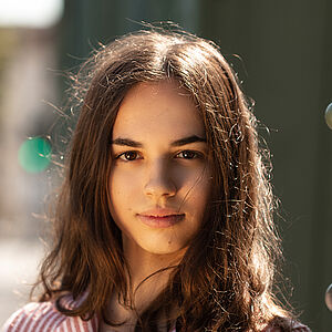 Portraitfoto von Veronica Scanferla. Sie trägt eine rot-weiß gestreifte Bluse mit einer aufgesetzten Tasche. Ihr braunes Haar liegt auf ihrer Schulter. Ihre rechte Hand liegt auf ihrer Brust.