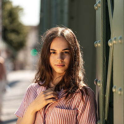 Portraitfoto von Veronica Scanferla. Sie trägt eine rot-weiß gestreifte Bluse mit einer aufgesetzten Tasche. Ihr braunes Haar liegt auf ihrer Schulter. Ihre rechte Hand liegt auf ihrer Brust.