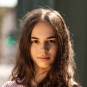 Portraitfoto von Veronica Scanferla. Sie trägt eine rot-weiß gestreifte Bluse mit einer aufgesetzten Tasche. Ihr braunes Haar liegt auf ihrer Schulter. Ihre rechte Hand liegt auf ihrer Brust.