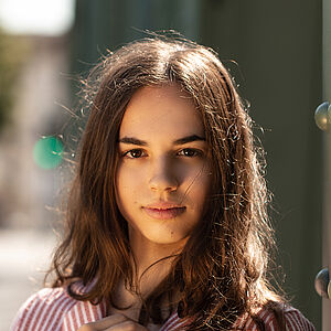 Portraitfoto von Veronica Scanferla. Sie trägt eine rot-weiß gestreifte Bluse mit einer aufgesetzten Tasche. Ihr braunes Haar liegt auf ihrer Schulter. Ihre rechte Hand liegt auf ihrer Brust.