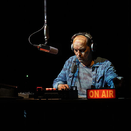 Ein Mann mit Kopfhörern auf dem Kopf sitzt an einem Tisch. Ein Mikrofon hängt von der Decke und Laptop und Mischpult stehen auf dem Tisch. Ein kleines Leuchtreklame Schild mit «ON AIR» darauf steht vor ihm.