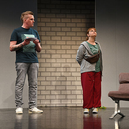 Tom Müller und Marie Petersen stehen auf der Bühne. Tom hat einen Zettel in der Hand und schaut entgeistert auf Marie. Er trägt ein blaues Shirt und eine graue Jeans. Marie ist von ihm abgewendet und starrt Löcher in die Luft.  Sie trägt eine rote Hose, gelbe Chucks und einen grauen Hoodie.