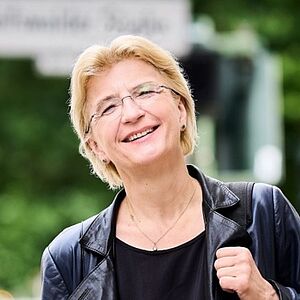 Portraitfoto von Antje Weithaas. Sie ist auf einer Straße unterwegs. Ihr Violinenkasten hängt über ihrer linken Schulter. Sie hält den Riemen in der Hand. Die linke Hand ist in ihrer Jackentasche. Sie trägt eine geöffnete schwarze Lederjacke, darunter ein schwarzes Oberteil. Ihr Kopf ist leicht schräg gelegt. Sie lächelt. Sie trägt eine randlose Brille und kurzes blondes Haar.