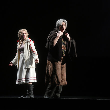 Solveig und Peer Gynts Mutter Åse sind auf der Bühne zu sehen.  Nur sie sind zu sehen der Rest ist in Dunkelheit gehüllt. Solveig trägt eine weiße Strickjacke und einen weißen Rock, jeweils mit roten Farbakzenten. Sie steht hinter Åse und schaut über ihre Schulter fragend zur ihr. Åse steht im Vordergrund. Sie trägt eine Jacke über ihren Schultern und singt, dem Publikum zugewandt.