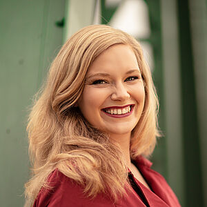 Portraitfoto von Signe Heiberg. Sie trägt ihr blondes Haar offen. Dazu roter Lippenstift und ein beerenroter Blazer. Sie lächelt mit geöffnetem Mund.