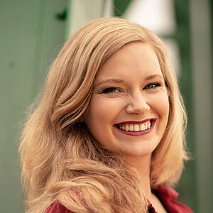 Portraitfoto von Signe Heiberg. Sie trägt ihr blondes Haar offen. Dazu roter Lippenstift und ein beerenroter Blazer. Sie lächelt mit geöffnetem Mund.