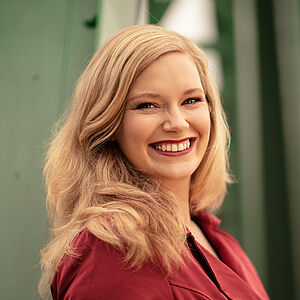 Portraitfoto von Signe Heiberg. Sie trägt ihr blondes Haar offen. Dazu roter Lippenstift und ein beerenroter Blazer. Sie lächelt mit geöffnetem Mund.