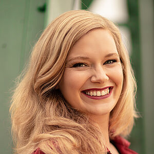 Portraitfoto von Signe Heiberg. Sie trägt ihr blondes Haar offen. Dazu roter Lippenstift und ein beerenroter Blazer. Sie lächelt mit geöffnetem Mund.