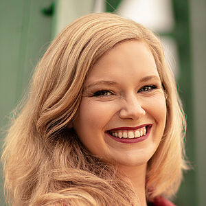 Portraitfoto von Victoria Kunze. Sie hat den rechten Arm um eine graue Säule gelegt, ihr Kopf lehnt daran. Sie trägt eine dunkelgrüne Bluse, darüber eine schwarze Jacke. Ihre blonden Wellen legen sich auf ihr Schultern.  