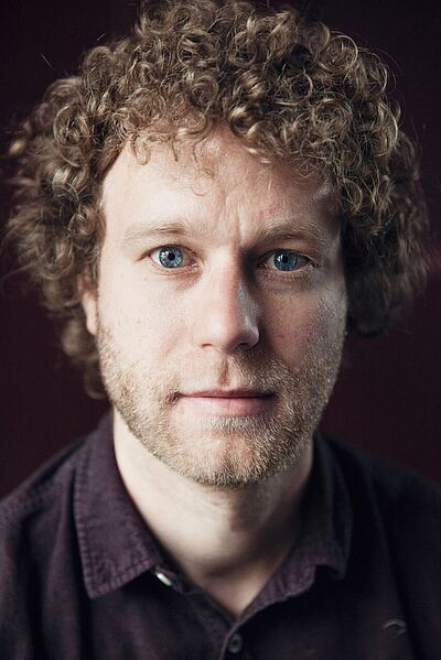 Portraitfoto von Wolfgang Nägele. Er hat lockiges dunkelblondes Haar, das sein Gesicht umrahmt sowie einen Dreitagebart. Seine hellblauen Augen stechen hervor. Er trägt ein violettes Hemd vor violettem Hintergrund.