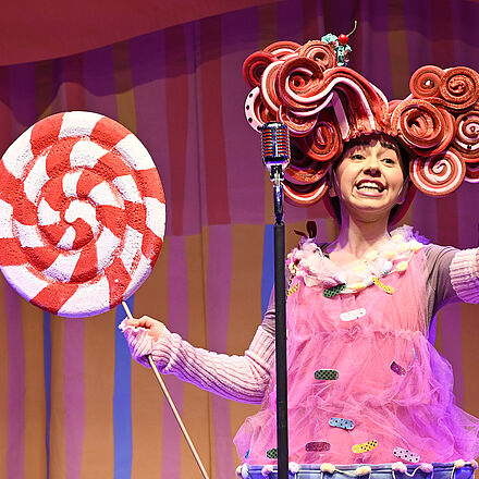 Im Mittelpunkt steht eine Frau im rosa Kleid mit einer roten Perücke auf dem Kopf und einem großen Lutscher in der Hand. Vor ihr steht ein Mikrofon.