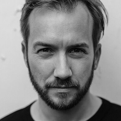 Schwarz-weißes Portraitfoto von Michael Müller-Kasztelan. Er steht mit kurzem Haar und Bart in einem schwaarzen T-Shirt und schaut mit starrem Blick.