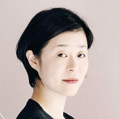 Portraitfoto von Mikiko Arai. Sie steht seitlich da und blickt in die Kamera. Sie hat schwarzes kurzes Haar und trägt ein schwarzes Oberteil.