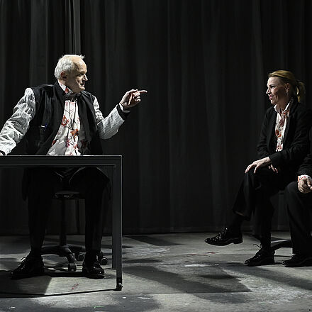 Links im Bild sitzt ein Mann an einem Tisch. Er schaut und zeigt nach rechts. Rechts im Bild sitzen zwei Personen, die den Mann ansehen.