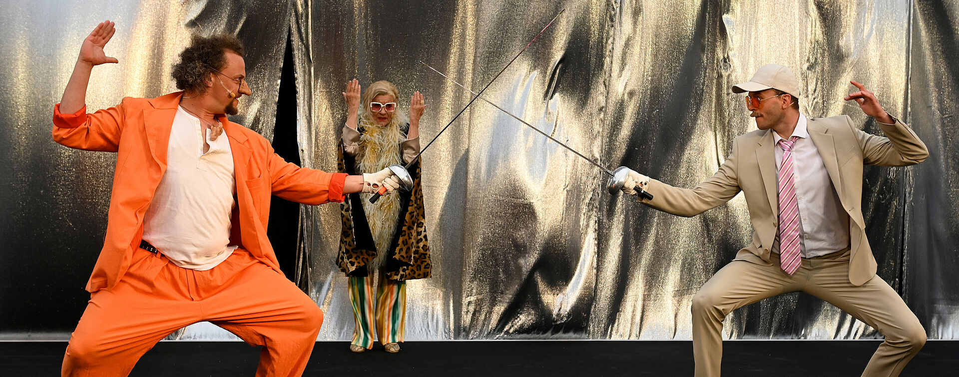 Cyrano und Valvert stehen sich gegenüber und kreuzen die Klingen ihrer Degen. Beide sind bereit für den ersten Streich. Im Hintergrund gibt eine Richterin den Startschuss. 