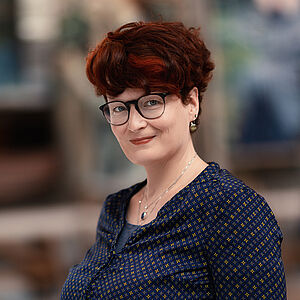 Portraitfoto von Bianca Sue Henne. Sie hat rotes lockiges kurzes Haar und trägt eine Brille. Sie steht seitlich und grinst leicht. Der Hintergrund ist verschwommen.