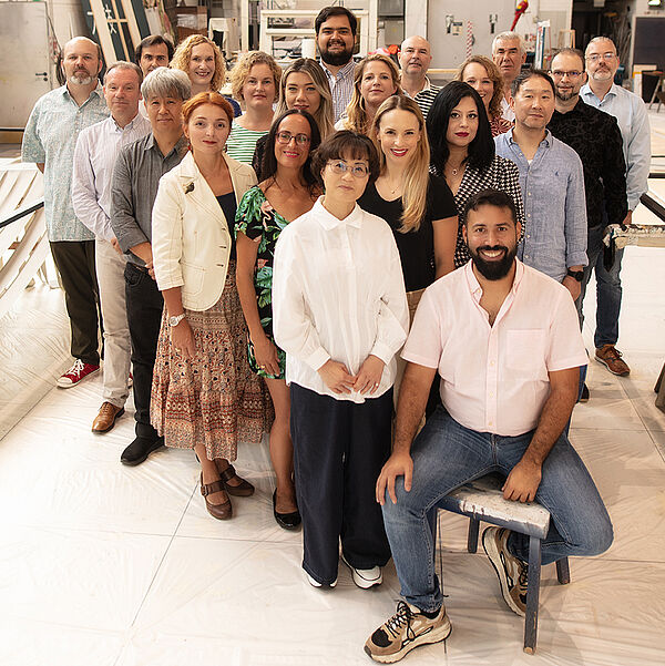 Fotos des Opernchors des Stadttheaters Bremerhaven. Sie sind in dreieckiger Formation aufgestellt. Chorleiter Mario El Fakih Hernández sitzt an der Spitze.