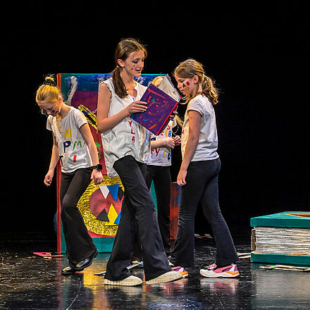 Vier Mädchen laufen auf der Bühne umher. Eines liest in einem Buch. Um sie herum liegen und stehen überdimensionierte Bücher. Sie tragen weiße T-Shirts mit bunten Buchstaben. Auf ihren Wangen sind bunte Buchstaben gemalt. 