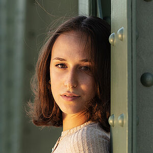 Portraitfoto von Deborah D’Aversa. Sie lehnt an einer grünen Metallstrebe und schaut mit gedrehtem Kopf nach vorn. Sie trägt ihr schulterlanges braunes Haar offen, dazu ein beigefarbenes geripptes Oberteil.