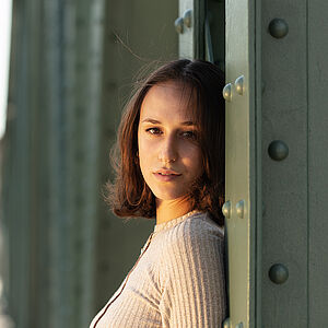 Portraitfoto von Deborah D’Aversa. Sie lehnt an einer grünen Stahlstrebe und schaut mit gedrehtem Kopf nach vorn. Sie trägt ihr schulterlanges braunes Haar offen, dazu ein beigefarbenes geripptes Oberteil.