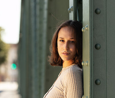 Portraitfoto von Deborah D’Aversa. Sie lehnt an einer grünen Stahlstrebe und schaut mit gedrehtem Kopf nach vorn. Sie trägt ihr schulterlanges braunes Haar offen, dazu ein beigefarbenes geripptes Oberteil.
