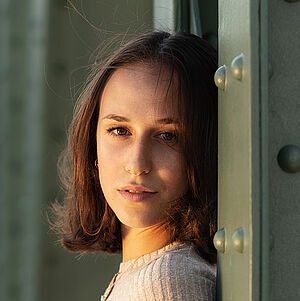 Portraitfoto von Deborah D’Aversa. Sie lehnt an einer grünen Metallstrebe und schaut mit gedrehtem Kopf nach vorn. Sie trägt ihr schulterlanges braunes Haar offen, dazu ein beigefarbenes geripptes Oberteil.