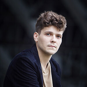 Portraitfoto von Szymon Nehring. Er trägt eine dunkelblaue Jacke über einem beigen Pullover. Sein dunkelblondes Haar ist locker nach oben gestylt. Er sitzt und blickt seitlich in die Kamera.
