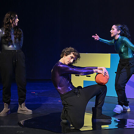 Auf dem Boden kniet eine Person und hält den Ball. Im Hintergrund stehen die beiden anderen