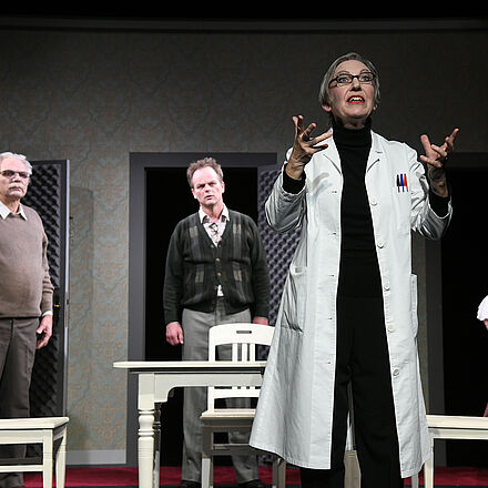 Im Vordergrund steht eine Frau mit einem Arztkittel, die die Arme nach vorne streckt. Im Hintergrund stehen drei Männer um einen Tisch herum. Der rechte Mann trägt Kleidung aus der Barock-Zeit.