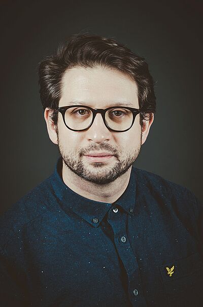 Portraitfoto von Sam Brown. Er trägt braunes Haar, eine eine dunkle Brille, einen Dreitagebart und ein dunkelblaues Hemd.