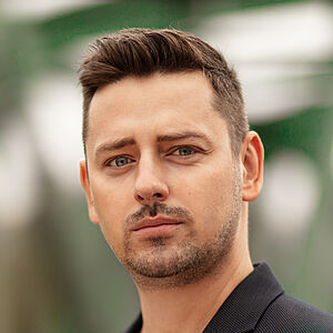 Portraitfoto von Marcin Hutek. Den Blick leicht in Richtung ferne schweifend. Er trägt er ein weißes Shirt unter einem schwarzen Jackett mit einem bunten Einstecktuch. 