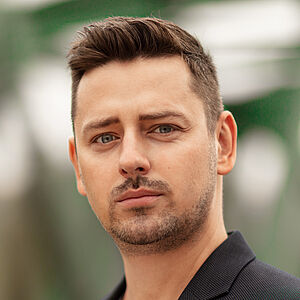 Portraitfoto von Marcin Hutek. Den Blick leicht in Richtung ferne schweifend. Er trägt er ein weißes Shirt unter einem schwarzen Jackett mit einem bunten Einstecktuch. 
