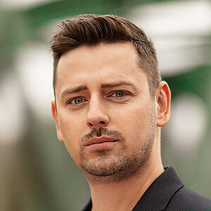 Portraitfoto von Marcin Hutek. Den Blick leicht in Richtung ferne schweifend. Er trägt er ein weißes Shirt unter einem schwarzen Jackett mit einem bunten Einstecktuch.
