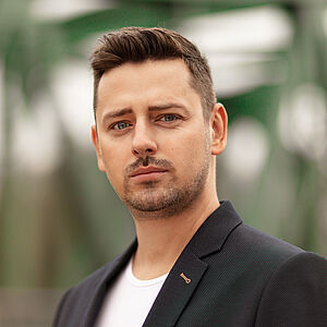 Portraitfoto von Marcin Hutek. Den Blick leicht in Richtung ferne schweifend. Er trägt er ein weißes Shirt unter einem schwarzen Jackett mit einem bunten Einstecktuch. 