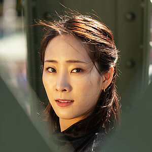 Portraitfoto von Yeojin Kim. Sie trägt ihr dunkles Haar halboffen. Es weht im Wind. Sie trägt ein schwarzes Oberteil. Sie blickt zwischen grünen Brückenstreben hindurch.