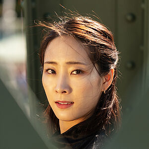 Portraitfoto von Yeojin Kim. Sie trägt ihr dunkles Haar halboffen. Es weht im Wind. Sie trägt ein schwarzes Oberteil. Sie blickt zwischen grünen Brückenstreben hindurch.