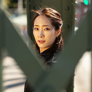 Portraitfoto von Yeojin Kim. Sie trägt ihr dunkles Haar halboffen. Es weht im Wind. Sie trägt ein schwarzes Oberteil. Sie blickt zwischen grünen Brückenstreben hindurch.