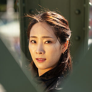 Portraitfoto von Yeojin Kim. Sie trägt ihr dunkles Haar halboffen. Es weht im Wind. Sie trägt ein schwarzes Oberteil. Sie blickt zwischen grünen Brückenstreben hindurch.