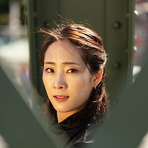 Portraitfoto von Yeojin Kim. Sie trägt ihr dunkles Haar halboffen. Es weht im Wind. Sie trägt ein schwarzes Oberteil. Sie blickt zwischen grünen Brückenstreben hindurch.