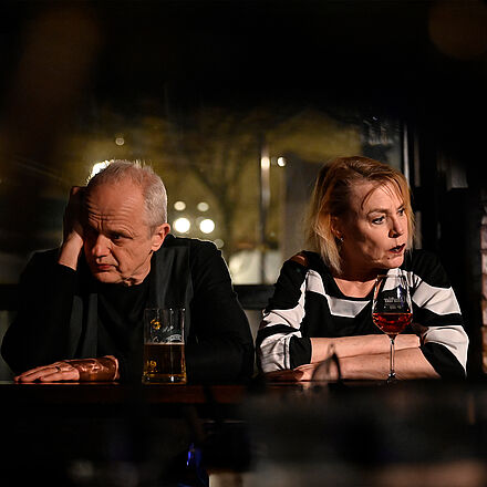 Eine Frau und ein Mann sitzen an einer Bar. Sie schauen weg. Vor ihnen stehen ein Glas Wein und ein Bier.