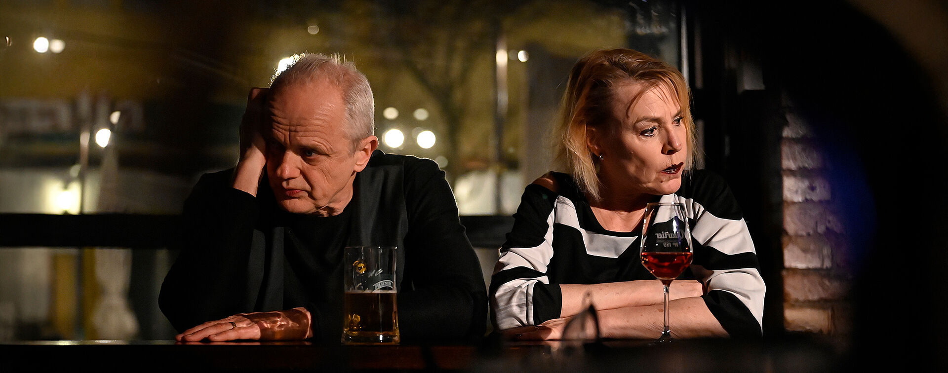 Eine Frau und ein Mann sitzen an einer Bar. Sie schauen weg. Vor ihnen stehen ein Glas Wein und ein Bier.