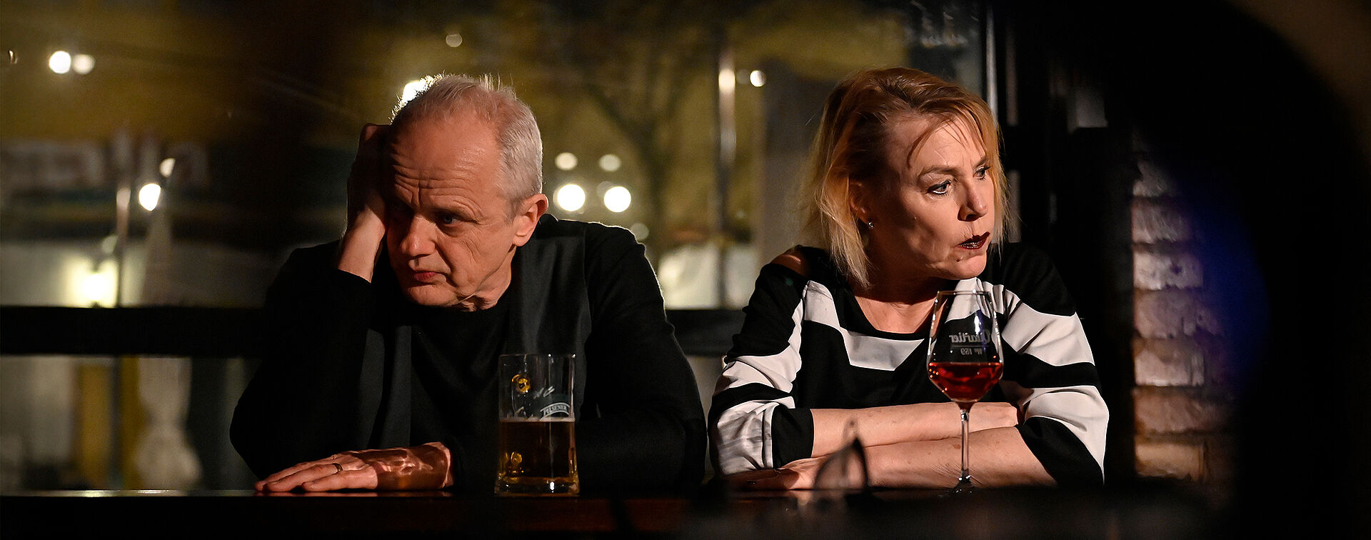 Eine Frau und ein Mann sitzen an einer Bar. Sie schauen weg. Vor ihnen stehen ein Glas Wein und ein Bier.