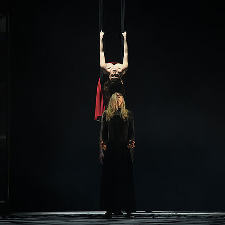 Eine Frau hängt von der Decke. Vor ihr steht eine Tänzerin. Sie schauen sich an.