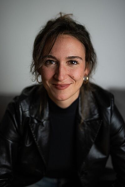 Portraitfoto von Ariane Königshof. Sie trägt eine schwarze Lederjacke übr einem schwarzen Pullover. Ihr Haar ist dunkelbraun und locker hochgesteckt. Sie grinst.