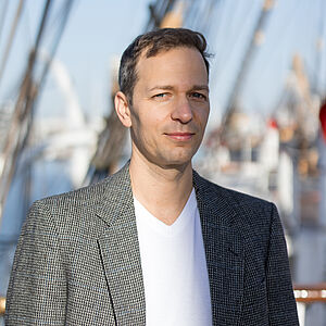 Portraitfoto von Balázs Szabó. Er steht leicht seitlich und blickt in die Kamera. Er ist in einen schwarz-weiß karierten Anzug gekleidet, darunter ein weißes Oberteil mit V-Ausschnitt. Die eine Hälfte seines Oberkörpers steht im Schatten, die andere in der Sonne. Im Hintergrund sind verschwommen die Seile und Taue eines Schiffes zu erkennen. 