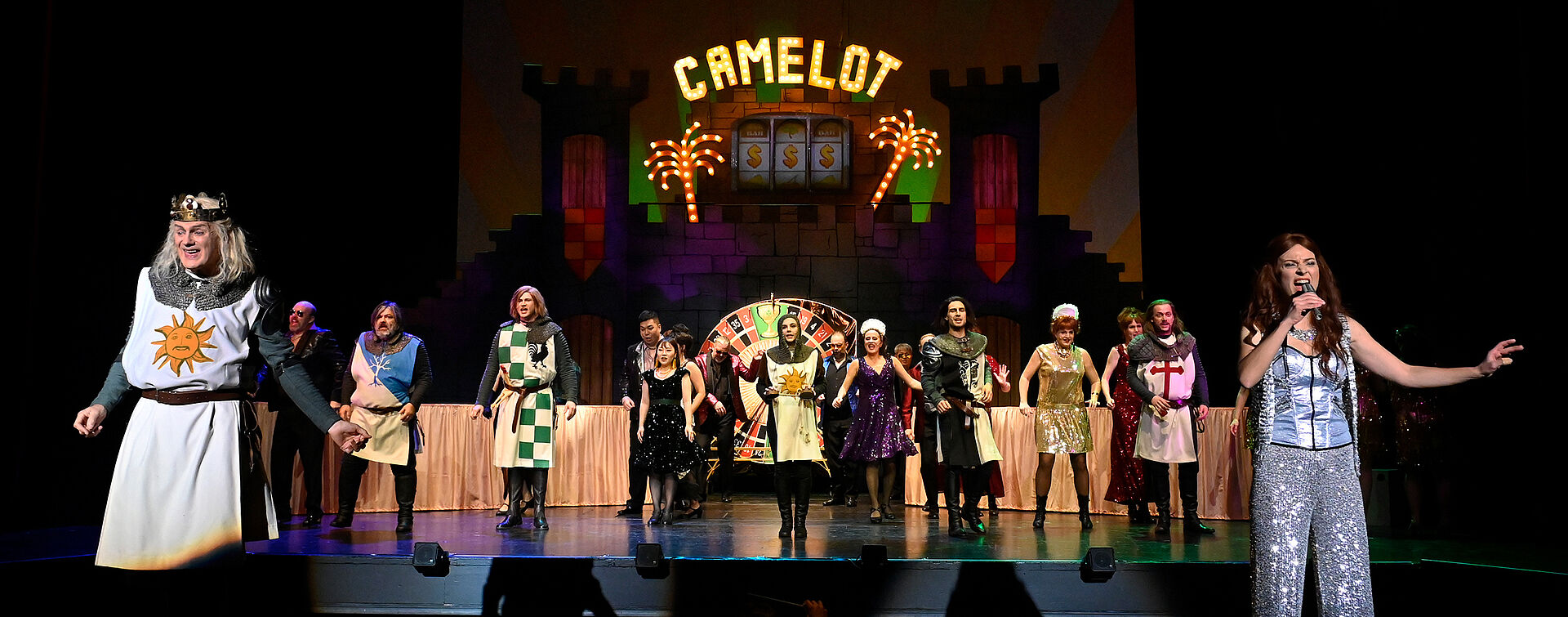Am vorderen Bühnenrand stehen König Artus und eine singende Frau in glitzerndem Kostüm. Im Hintergrund tanzt eine Gesellschaft vor der Burg Camelot, deren Name in leuchtenden Lettern erstrahlt.