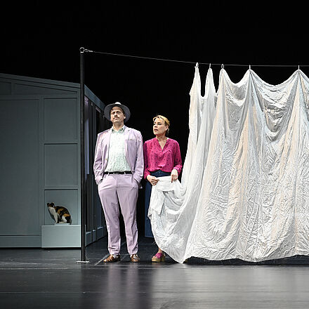 Eine Frau hält eine Wäscheleine in der Hand. Neben ihr steht ein Mann mit Händen in den Taschen und Hut. Im Hintergrund erkennt man ein Haus und eine Katze.