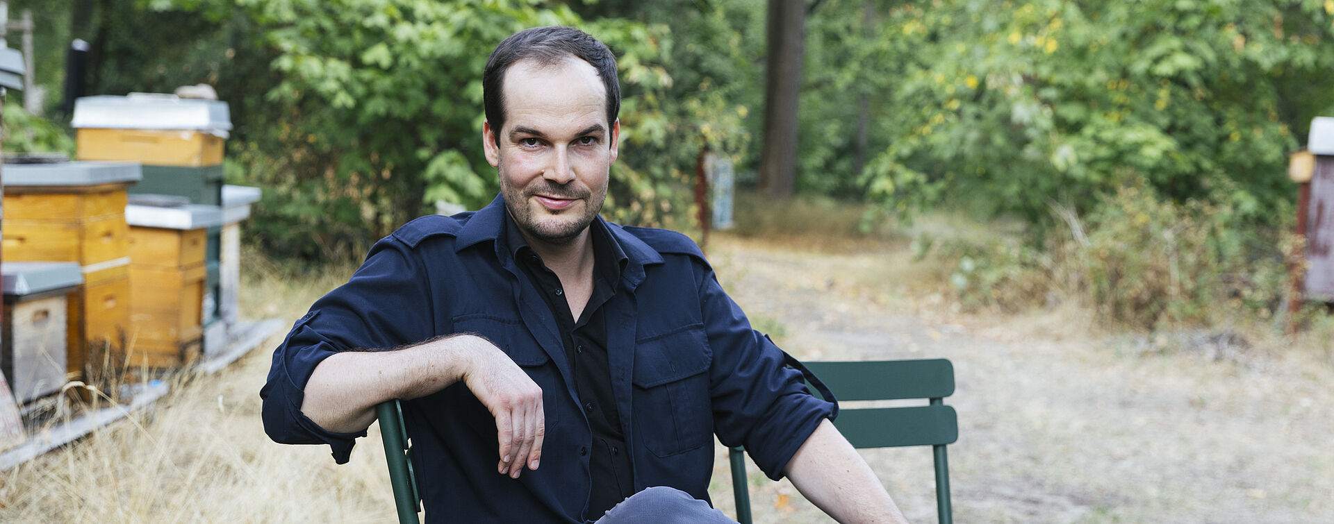 Portraitfoto von Max Czollek. Er sitzt seitlich auf einer dunkelgrünen Bank. Er hat dunkles Haar und einen Dreitagebart. Er trägt ein dunkelblaues Hemd über einem schwarzen T-Shirt. Hinter ihm erstreckt sich eine Wiese auf der Bienenkörbe stehen. 